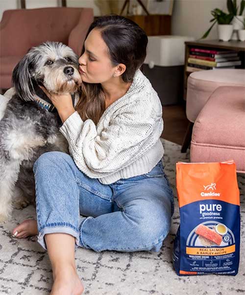 Young Woman sitting on the floor and kissing her dog, with bag of Canidae Pure dog food next to her.