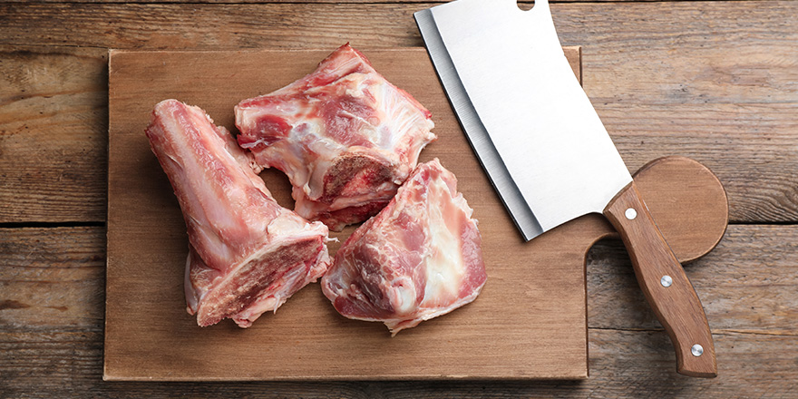Cutting board with raw chopped meaty bones and butcher knife on wooden table, top view