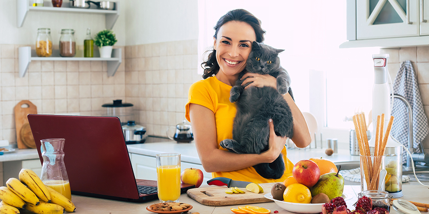 Cute beautiful and happy young brunette woman with her cat in the kitchen at home is preparing fruit vegan salad or a healthy smoothie and having fun