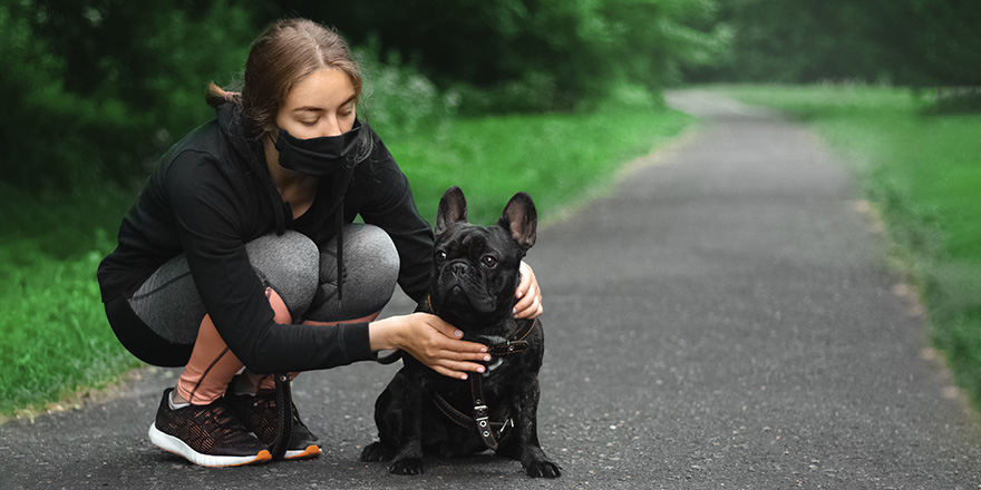 A young woman during pandemic isolation walking with dog in park wearing disposable medical face mask. 
