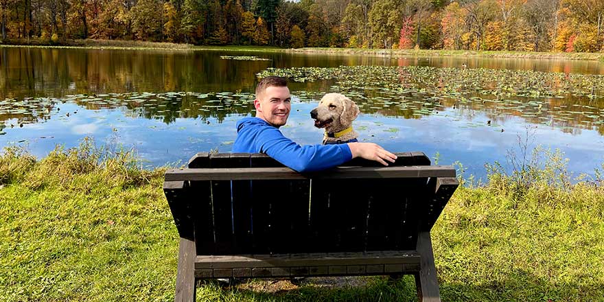 Young man sitting on a bench with a Goldendoodle dog in front of a lake and woods at Cuyahoga Valley National Park