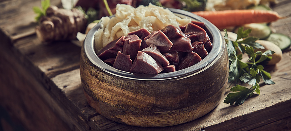 From above of raw cut meat and cartilages in dog bowl placed on lumber table near herbs and assorted vegetables