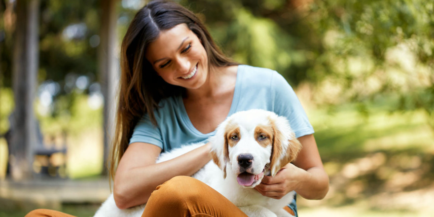 A young woman is sitting in the park with a white and brown puppy in her lap. 